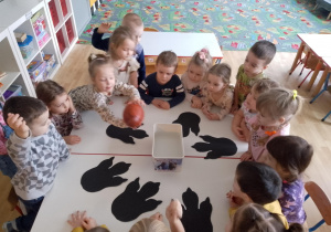 Sala przedszkolna. Dzieci przy stole oglądają ,jajo dinozaura" przed włożeniem do wody. Na stole rozłożone są ślady dinozaurów wycięte z papieru.