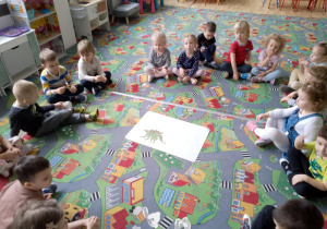 Sala przedszkolna. Dzieci siedzą na dywanie w kole. Na środku leży zdjęcie dinozaura.