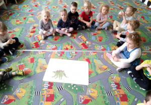 Sala przedszkolna. Dzieci siedzą na dywanie w kole. Na środku leży zdjęcie dinozaura.