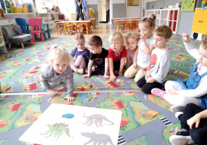 Sala przedszkolna. Dzieci siedzą na dywanie w kole. Na środku leżą zdjęcia dinozaurów. Dziewczynka układa kolejne zdjęcie dinozaura.