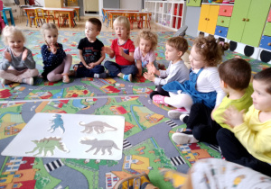 Sala przedszkolna. Dzieci siedzą na dywanie w kole. Na środku leżą zdjęcia dinozaurów.