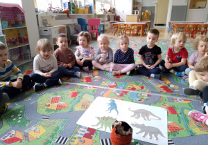 Sala przedszkolna. Dzieci siedzą na dywanie w kole. Na środku leżą zdjęcia dinozaurów i pęknięte ,,jajo dinozaura".