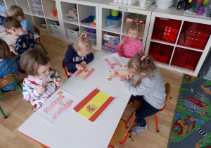 Sala przedszkolna. Dzieci siedzą przy stole i wykonują pracę plastyczną ,,Flaga Hiszpanii".