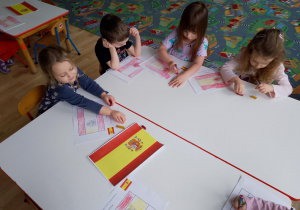 Sala przedszkolna. Dzieci siedzą przy stole i wykonują pracę plastyczną ,,Flaga Hiszpanii".
