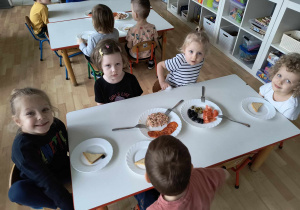 Sala przedszkolna. Dzieci siedzą przy stole próbują hiszpańskie produkty.