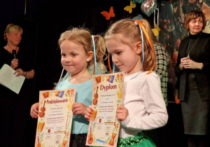 Dwie dziewczynki stojąc na scenie trzymają w dłoniach dyplomy