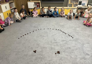 Dzieci w kole na dywanie układają uśmiechniętą minkę z kasztanów