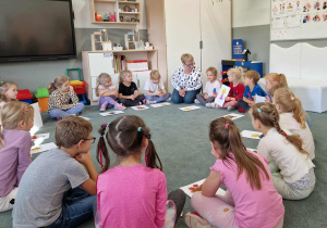 Dzieci siedzą w kole na dywanie i prezentują samodzielnie wykonane ludziki z jesiennych liści