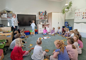 Dziewczynka pokazuje dzieciom układ ruchów, który dzieci, zgodnie z przyjętym kodem, odtworzą za chwilę wykorzystując leżące przed nimi dary jesieni