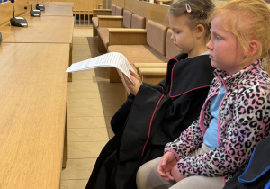Dziewczynki siedzą w ławie sądowej. Dziewczynka wcielająca się w rolę prokuratora czyta dokument aktu oskarżenia