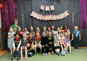 Dzieci z Grupy Sówki pozują wraz z nauczycielką do zdjęcia na tle dekoracji karnawałowej