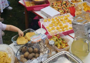 Ogród przedszkolny. Dzieci i rodzice na pikniku rodzinnym zgromadzeni przy stole, na którym znajdują się potrawy przygotowane przez rodziców.