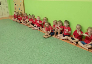 Dzieci w strojach gimnastycznych machają do zdjęcia siedząc w szeregu przy ścianie