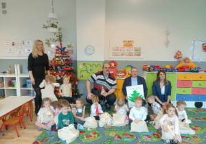 Sala przedszkolna. Dzieci z grupy ,,Wiewiórek", siedzą na dywanie, trzymają świąteczne paczki. Na zdjęciu z dziećmi siedzą przedstawiciele Rady Osiedla ,,Karolew" w Łodzi, pani dyrektor Przedszkola Miejskiego nr 112 w Łodzi oraz wychowawczyni grupy. W tle widoczna choinka.