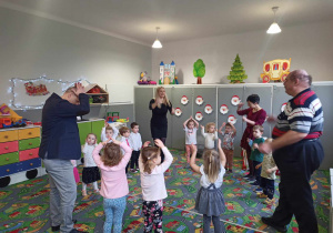 Sala przedszkolna. Dzieci z grupy ,,Wiewiórek" wraz z przedstawicielami Rady Osiedla ,,Karolew" w Łodzi oraz wychowawczynią grupy i pani z obsługi przedszkola, biorą udział w zabawie muzyczno-ruchowej.